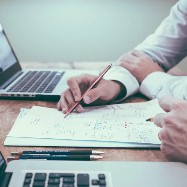 Two men sitting at a desk with laptops looking at some papers