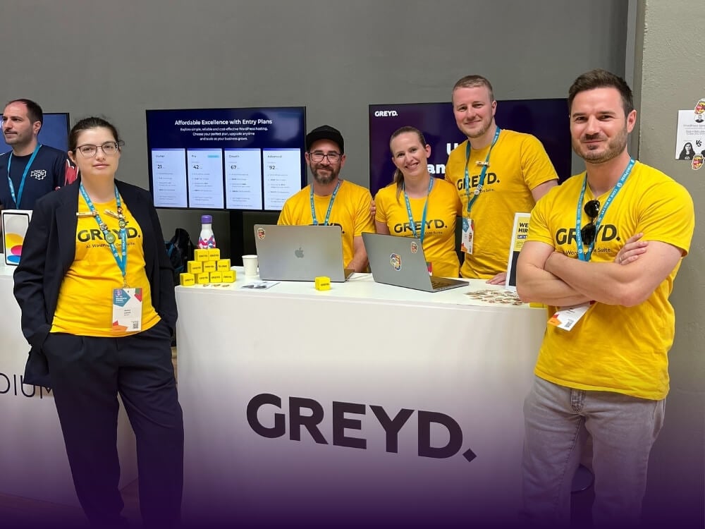 Five members of team Greyd, two women and three men, in yellow t-shirts around a white hostess desk with the Greyd logo.