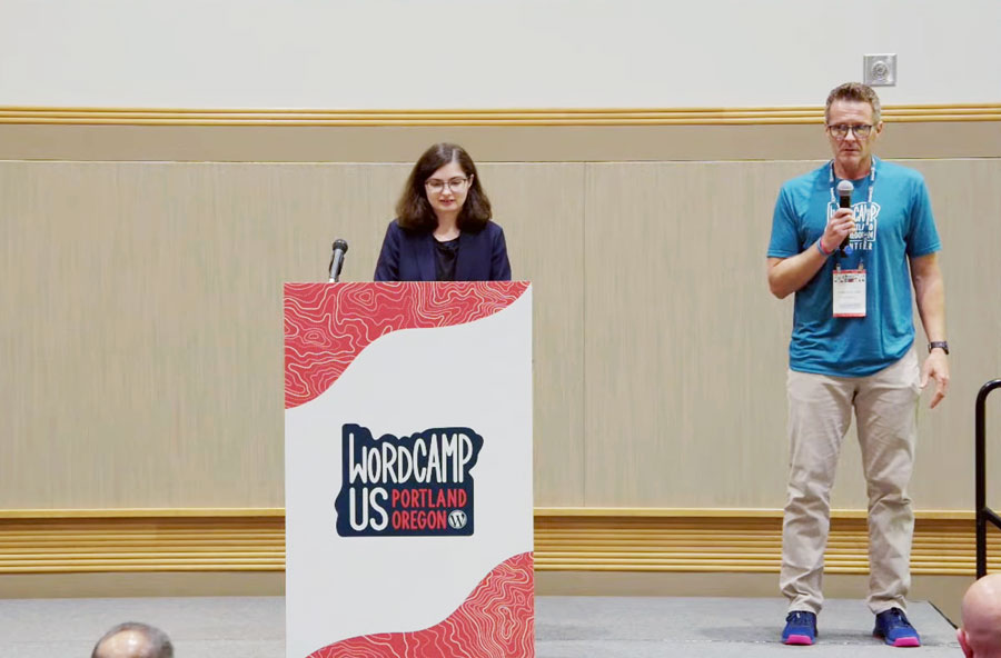 Jessica Lyschik standing behind a podium with the WordCamp US Portland Oregon 2024 logo, preparing to present, while a co-presenter stands beside her holding a microphone.