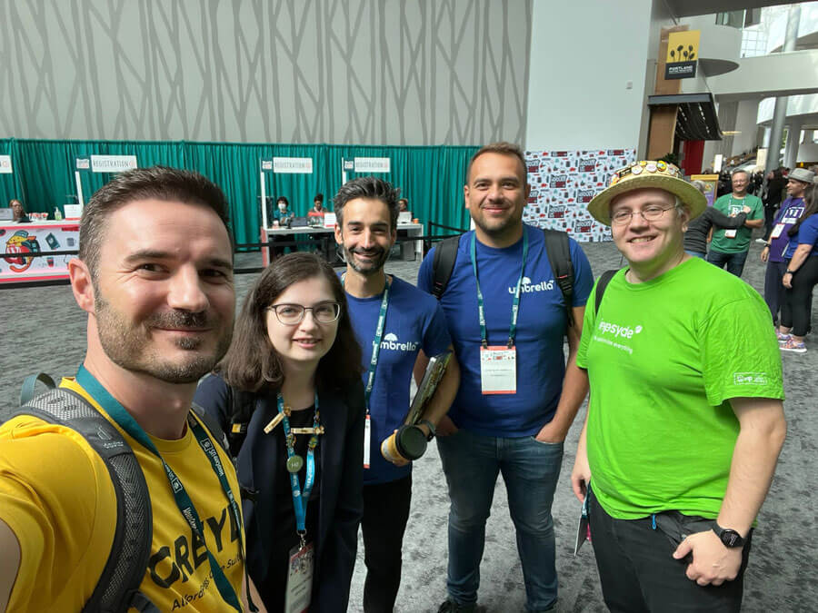 Mark Weisbrod, Jessica Lyschik, Aurelio Volle, and Robert Windisch smiling for a group photo at WordCamp US 2024, standing in front of the registration area with green curtains in the background.