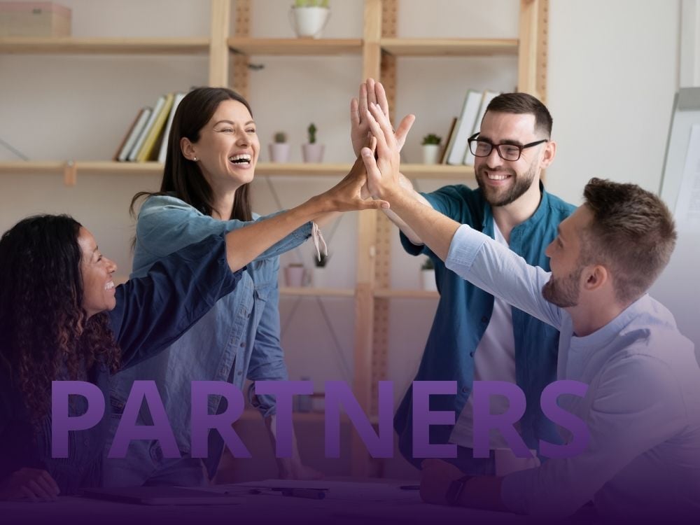 Group of four smiling colleagues high-fiving at a desk, symbolizing teamwork and collaboration, with the word “PARTNERS” overlaid in bold purple text.