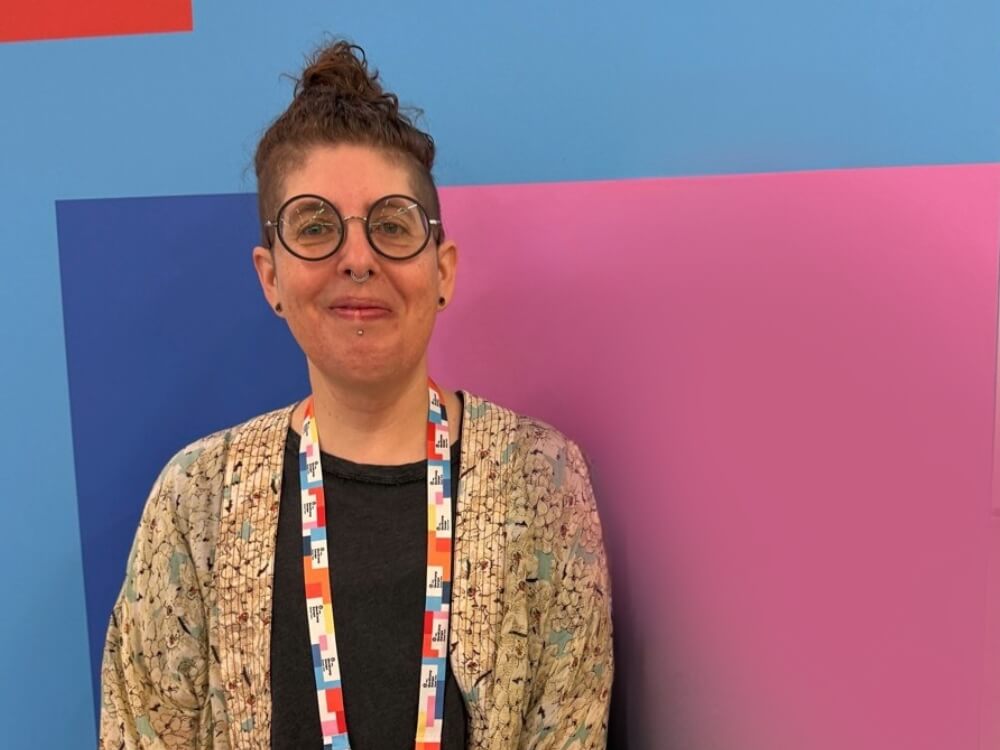 Tammie Lister, smiling, wearing round glasses and short curly hair in a top knot, wearing a patterned cardigan and colorful event lanyard, standing in front of a blue and pink geometric background.
