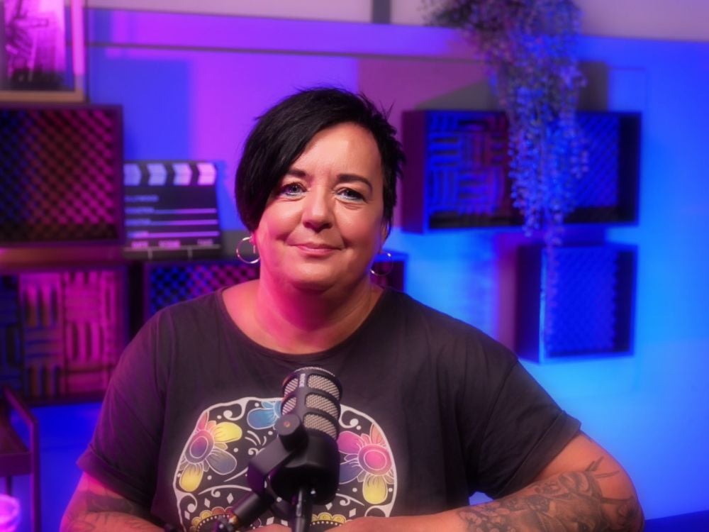 Woman with short dark hair and hoop earrings sitting in a recording studio, smiling at the camera. She wears a dark t-shirt with a colorful floral graphic and sits behind a microphone. The studio is lit with pink and blue lighting, with acoustic panels and film props in the background.