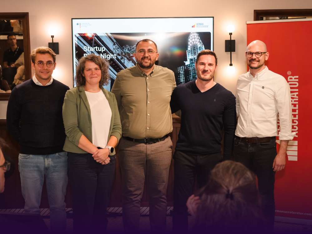Group photo of five participants from the German Accelerator program standing together after the Startup Pitch Night, with the event screen and banner visible in the background.