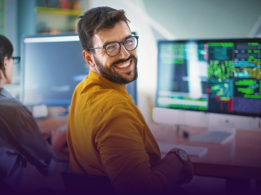 A smiling man with glasses and a beard sits in front of a computer screen displaying code, wearing a mustard-colored shirt and looking back at the camera.
