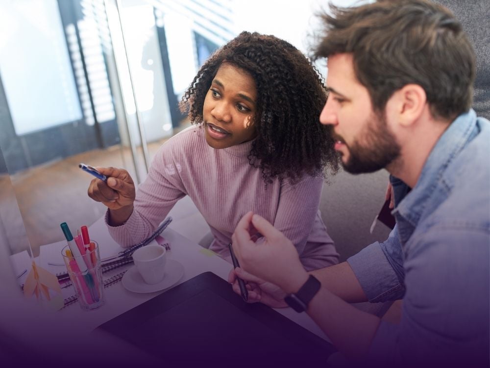 A woman with curly hair points with a pen while conversing with a bearded man at a desk with markers and a tablet.